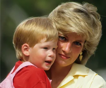 A woman in a yellow shirt lovingly holds a young child in a red outfit, with a blurred green background.