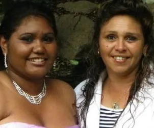 Two women smiling; the one on the left wears a strapless dress and jewelry, the one on the right wears a white jacket.