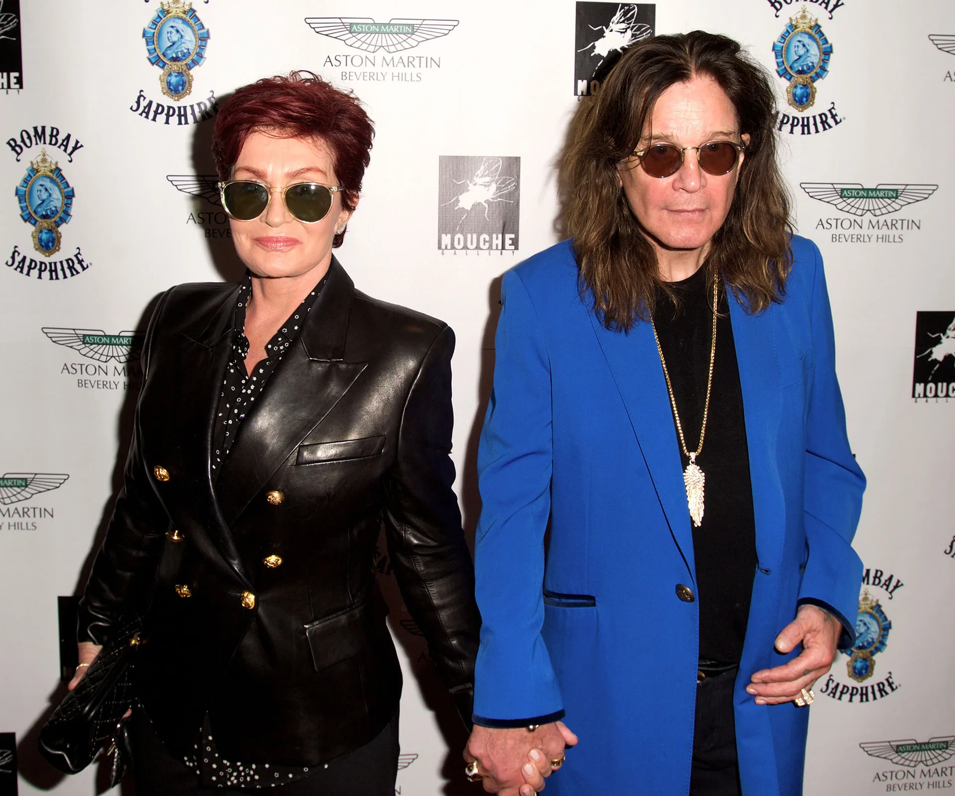 A couple holding hands, one in a black jacket and sunglasses, the other in a blue coat, on a red carpet event backdrop.