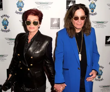 A couple holding hands, one in a black jacket and sunglasses, the other in a blue coat, on a red carpet event backdrop.
