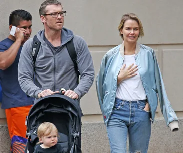 A man pushes a stroller with a child, while a woman walks beside him in casual clothing on a city street.