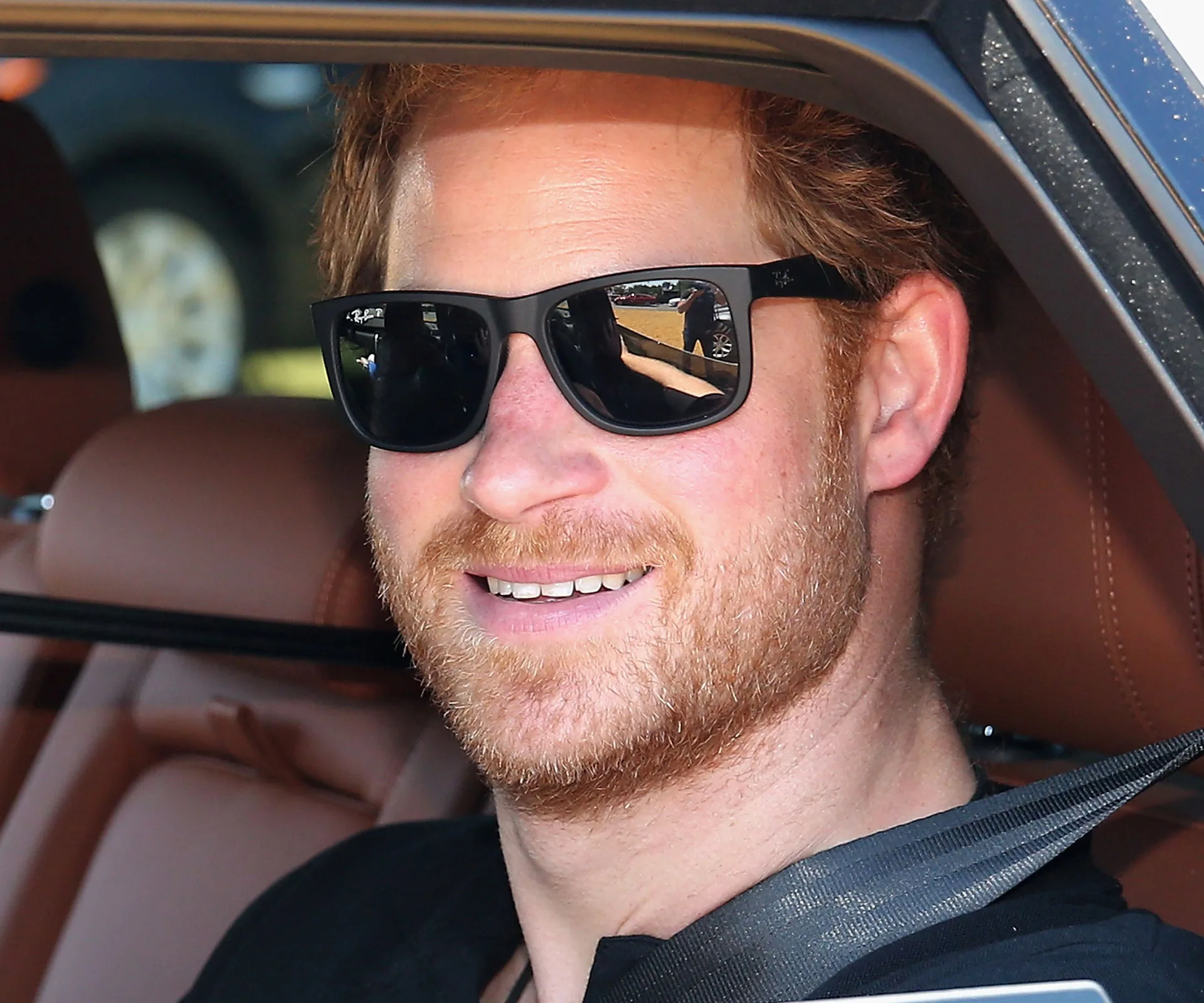 A man with red hair and sunglasses smiling while seated in a car.