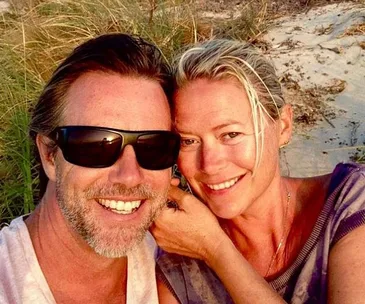 A happy couple posing together on a beach at sunset, with smiles and greenery in the background.
