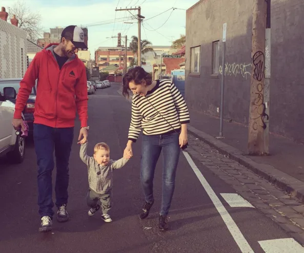 "Three people walking on a street; two adults holding hands with a joyful toddler between them."