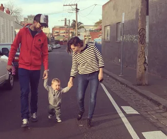 "Three people walking on a street; two adults holding hands with a joyful toddler between them."