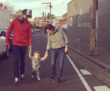 "Three people walking on a street; two adults holding hands with a joyful toddler between them."