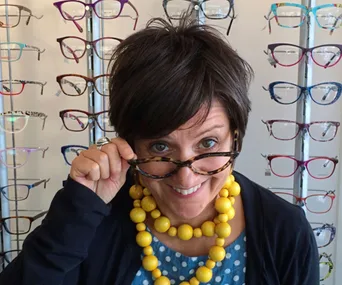 Woman smiling and holding glasses at an eyewear store with various frames displayed in the background.