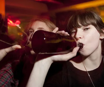 Two young people drinking from beer bottles in a dimly lit setting.