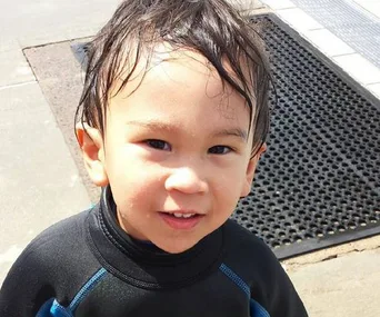 Young child with wet hair in a wetsuit, smiling outdoors.