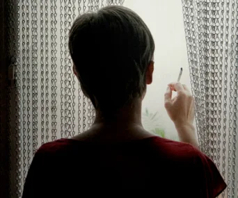 A person standing by a window, holding a cigarette, with light filtering through a chain curtain.