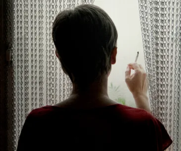 A person standing by a window, holding a cigarette, with light filtering through a chain curtain.