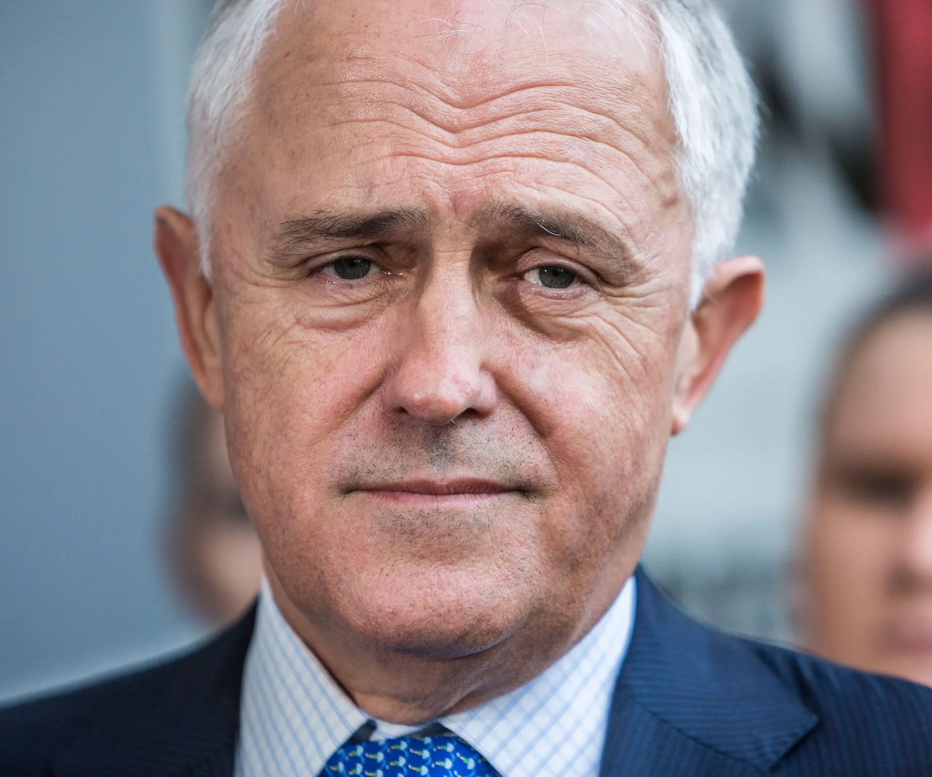 Portrait of an older man in a suit with a striped shirt and blue tie, showing a neutral expression.