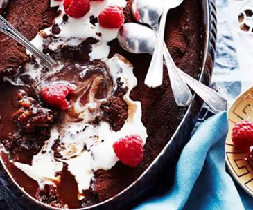 Chocolate pudding with cream, topped with raspberries, with spoons in a black dish on a blue cloth.