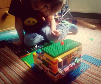 Child playing with a colorful Lego house model on a striped carpet indoors.