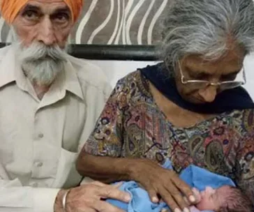 An elderly couple gently holding their newborn baby, wrapped in a blue blanket, with a patterned background.