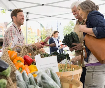 "Customers buying fresh produce from a smiling vendor at a farmers market stall under a white canopy."