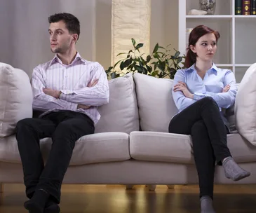 Couple sitting apart on a sofa, arms crossed and looking away, conveying tension.