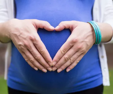 Pregnant woman in blue shirt forms heart shape with hands on belly, symbolizing love and hope.