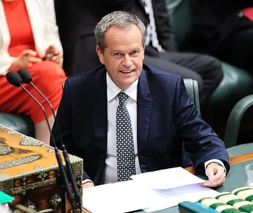 A man in a suit and patterned tie sits smiling at a desk, holding papers during a formal event or meeting.
