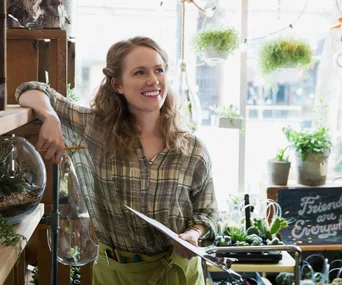 Young woman in a plaid shirt smiling while standing in a plant shop, holding a clipboard and pencil.