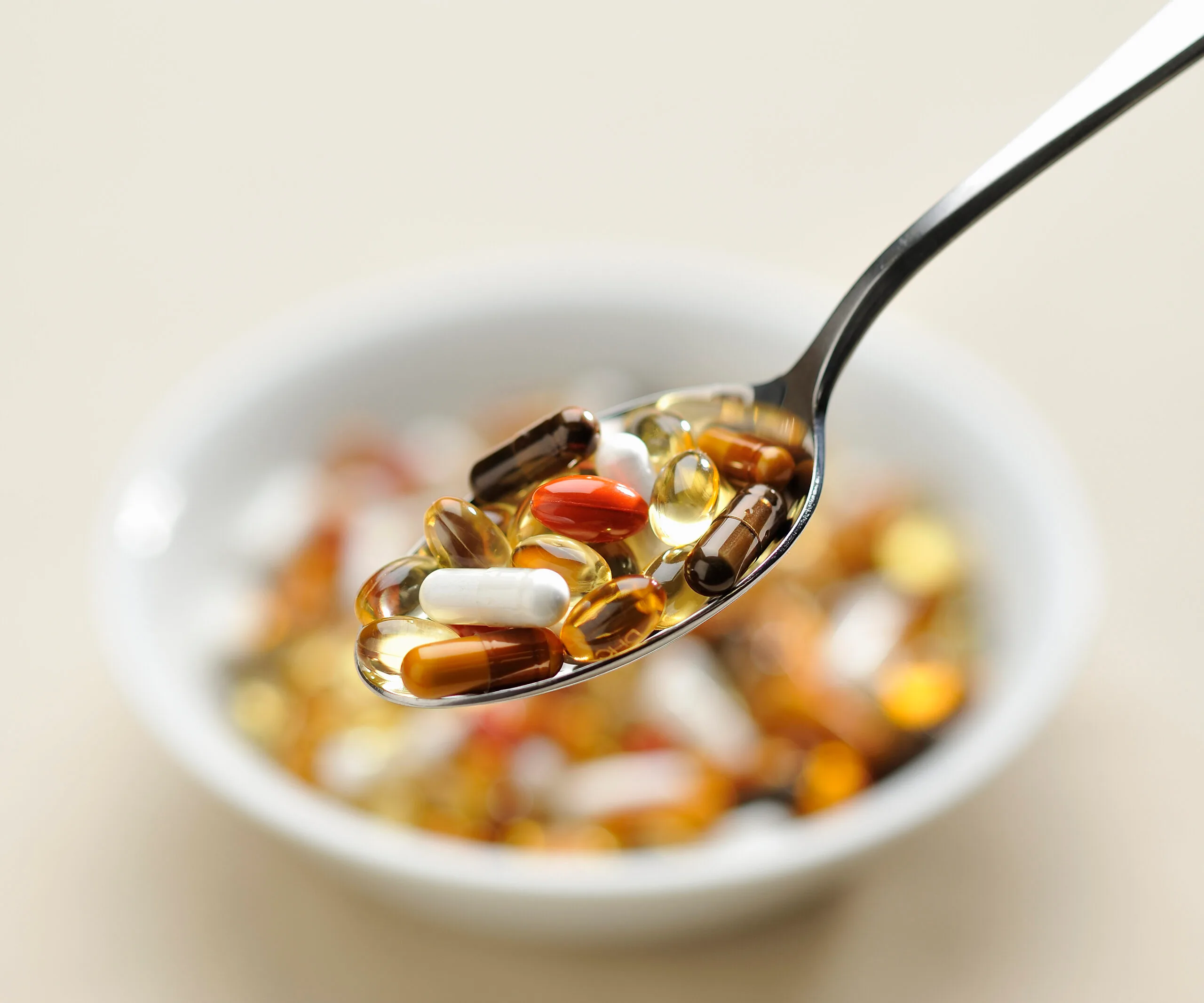 A spoonful of various colorful vitamin capsules and tablets above a bowl.