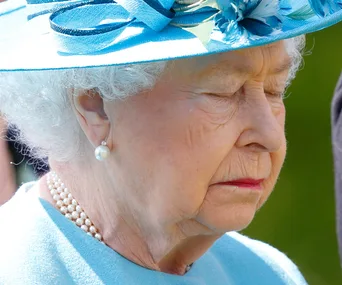 An elderly woman in a blue hat with flowers, eyes closed, wearing pearl earrings and necklace, outdoors.