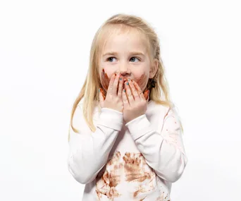A young girl with blonde hair, chocolate-covered hands and face, wearing a white shirt, looking away playfully.