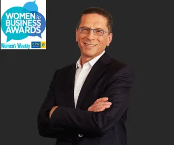 A person in a suit smiles with arms crossed next to "Women in Business Awards" logo on a dark background.