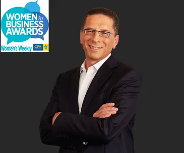 A person in a suit smiles with arms crossed next to "Women in Business Awards" logo on a dark background.