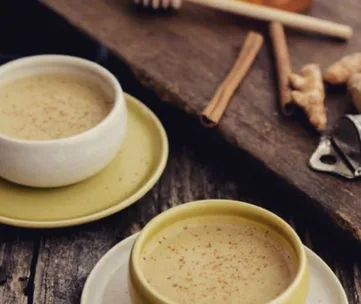 Two cups of golden milk on saucers with cinnamon sticks and ginger on a rustic wooden surface.
