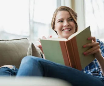 Woman smiling while sitting on a couch, holding an open book.