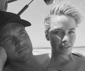A couple posing on a beach in black and white, the woman smiling and the man wearing a cap, looking thoughtful.