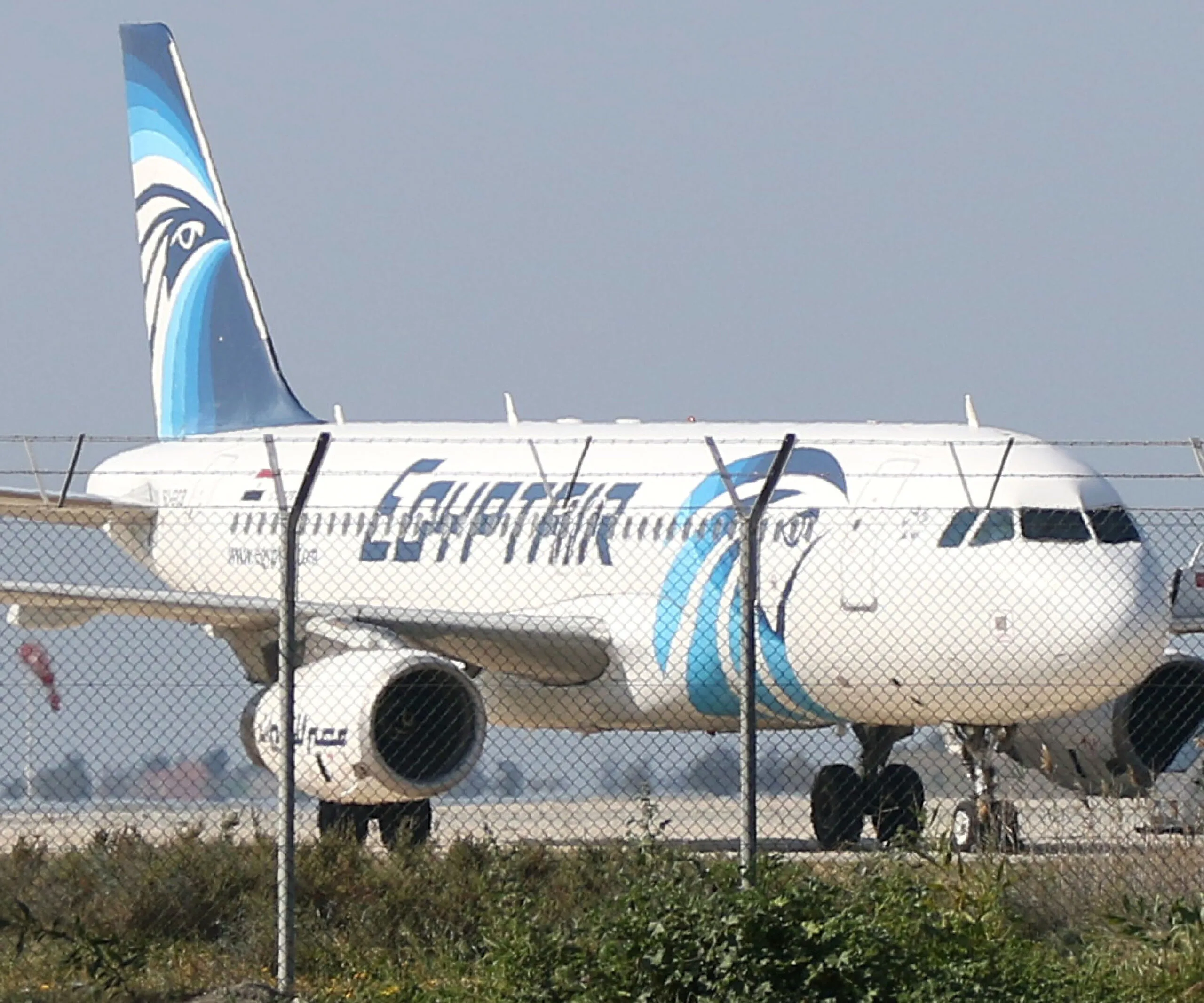 EgyptAir airplane on runway behind a fence, with the airline's logo visible on the tail and fuselage.