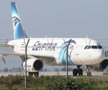 EgyptAir airplane on runway behind a fence, with the airline's logo visible on the tail and fuselage.