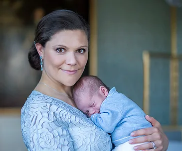 Mother in blue dress holding a sleeping baby in a light blue outfit.