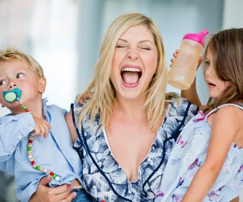 A mother laughs while holding a young boy with a pacifier and a girl with a sippy cup.