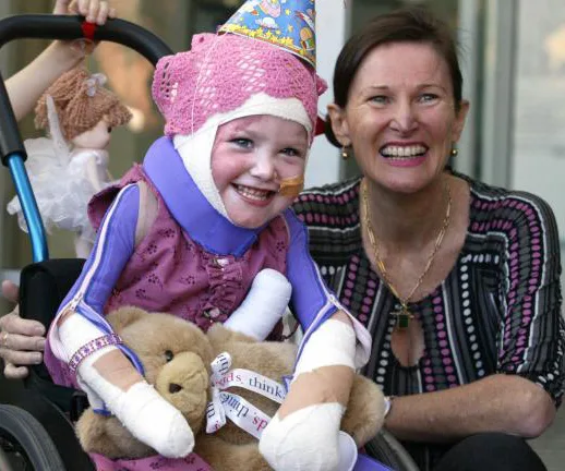 Girl in a wheelchair wearing a party hat, bandages, and holding a teddy bear, smiles with a woman beside her.