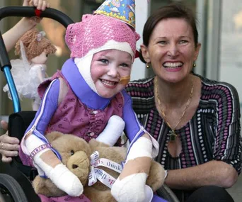 Girl in a wheelchair wearing a party hat, bandages, and holding a teddy bear, smiles with a woman beside her.
