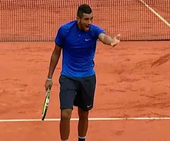 A tennis player in a blue shirt passionately gestures on a clay court during a match.