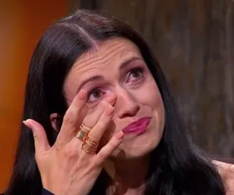 Woman with long dark hair and rings wiping a tear from her eye, appearing emotional.