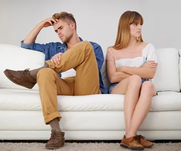 A couple sitting on a white couch looking upset and facing away from each other, suggesting a disagreement or tension.