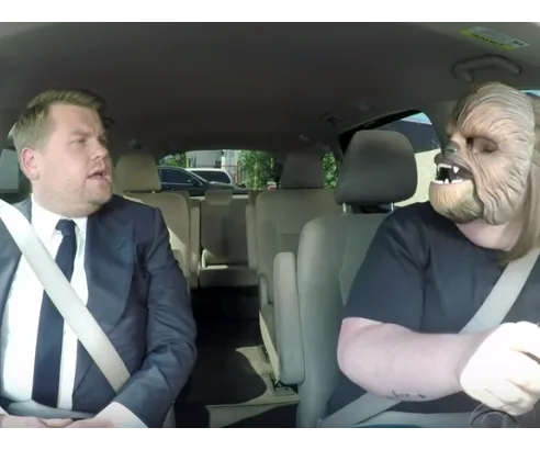 James Corden in a car with a person wearing a Chewbacca mask, both in seatbelts.