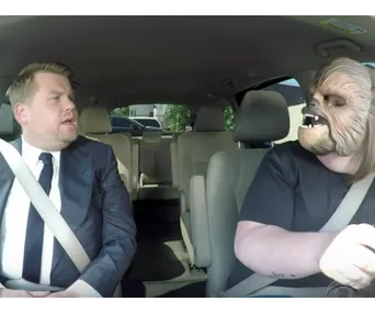 James Corden in a car with a person wearing a Chewbacca mask, both in seatbelts.