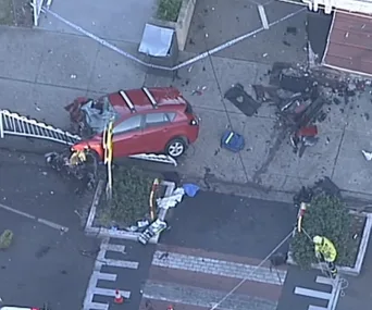 Red car crashed into a barrier on a sidewalk, debris scattered, emergency tape present.