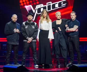 Judges of "The Voice" stand on stage, posing confidently with the show's logo in the background.