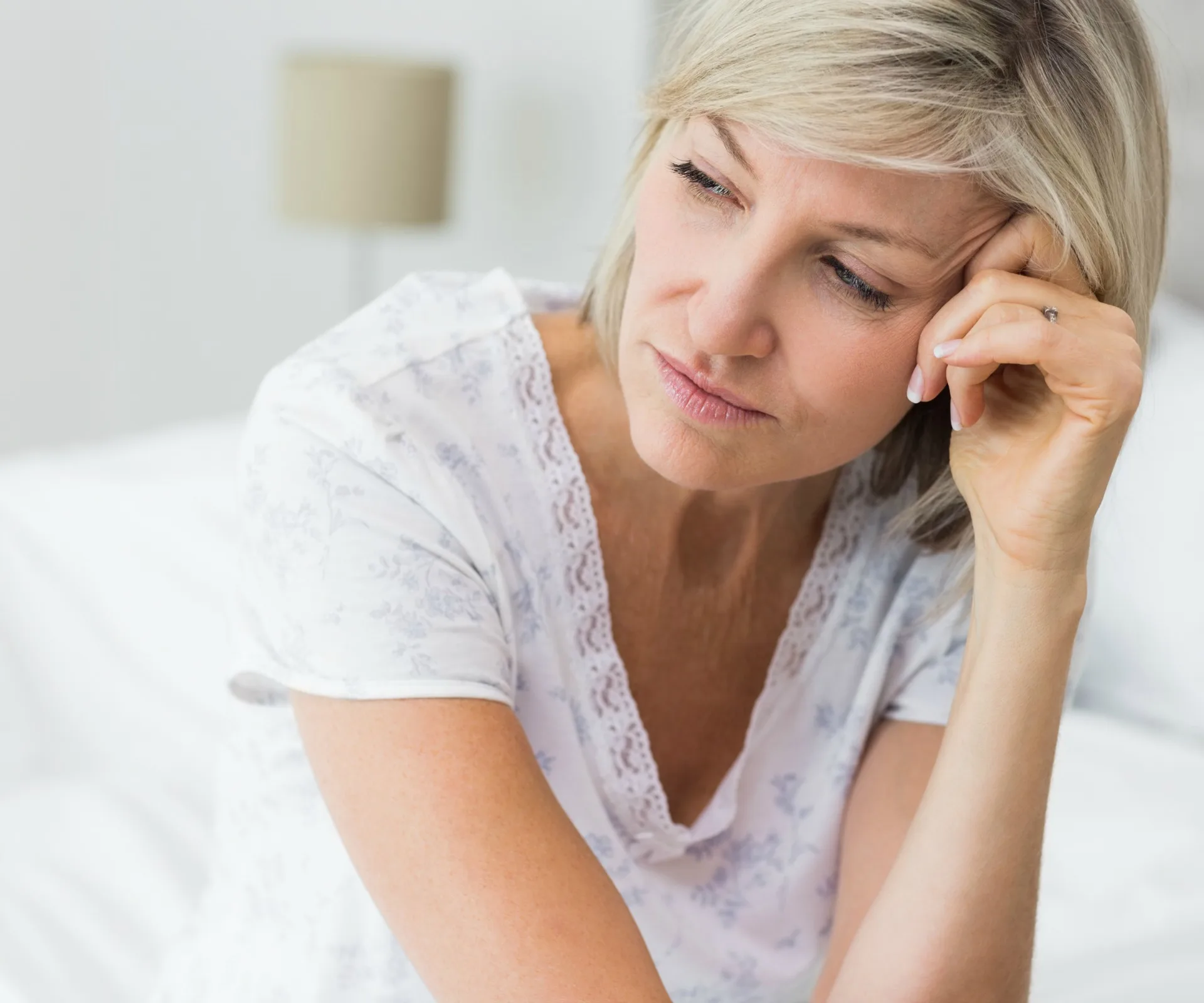 A woman with short blonde hair looks pensive, resting her head on her hand, sitting on a bed.