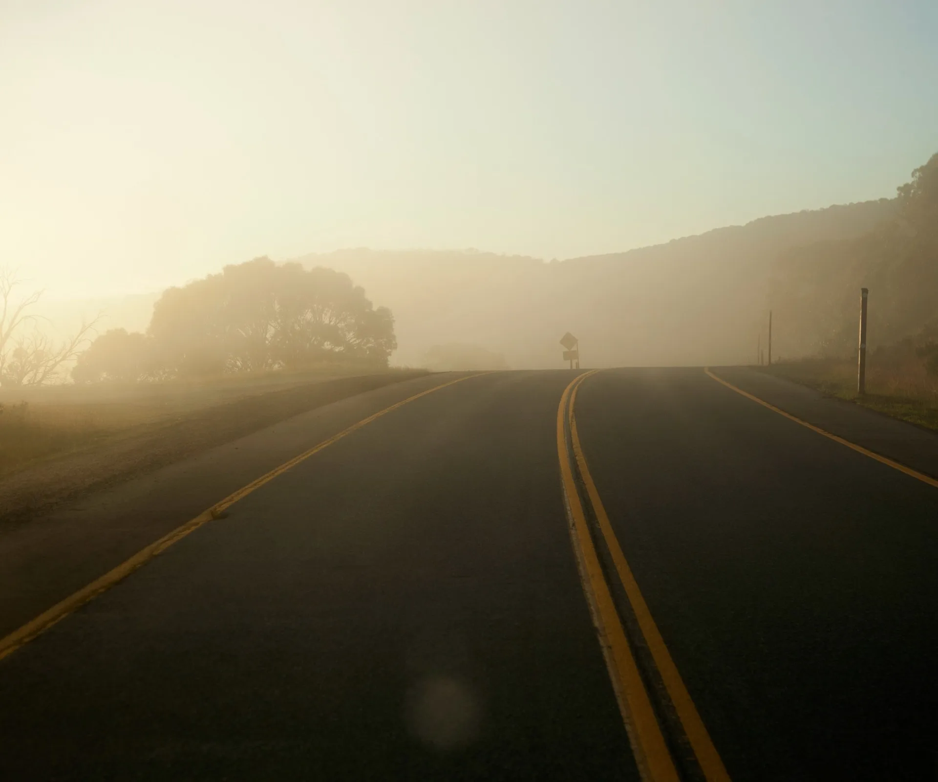 Foggy scenic road with trees and hills in the distance under a soft, hazy sky.
