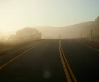 Foggy scenic road with trees and hills in the distance under a soft, hazy sky.
