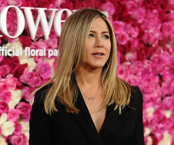 A woman with long blonde hair in a black outfit stands in front of a wall of pink roses.