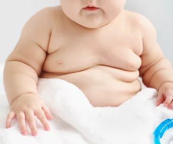 Plump baby girl sitting on a white towel, showing chubby arms and belly.